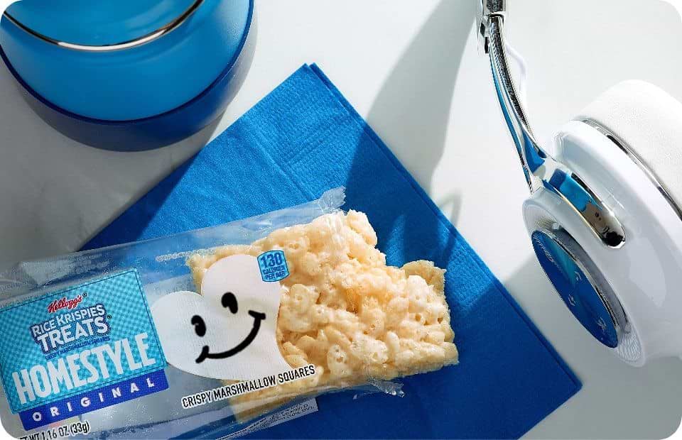 An opened and bitten Rice Krispies Treats® homestyle bar on a blue napkin with a cup of water and white headphones
